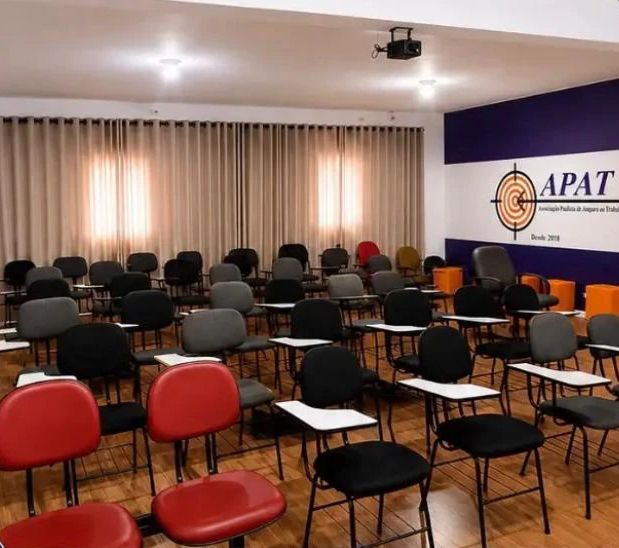 Auditório da APAT com cadeiras com pranchetas, cortinas e o painel com o logotipo da APAT ao fundo.