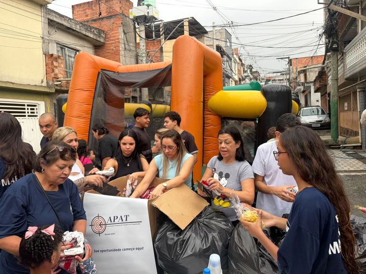 Equipe da APAT separando doces e brinquedos ao lado da faixa "Sua oportunidade começa aqui!", com um brinquedo inflável ao fundo.