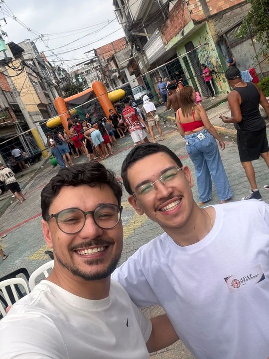 Participantes sorrindo em uma selfie, com a festa e o brinquedo inflável ao fundo.